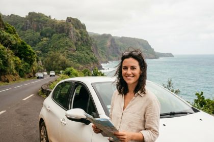 Femme souriante appuyée contre une voiture de location sur une route côtière de La Réunion avec vue sur l'océan Indien
